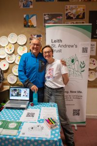 Trustees Daniel and Nina at a Leith St. Andrew's Trust stall. They're facing the camera, with Daniel wearing a blue jumper, Nina wearing a white t-shirt. Behind them is a green and white Leith St. Andrew's Trust pop-up banner and in front of them is a table with flyers and a laptop with info about the project.